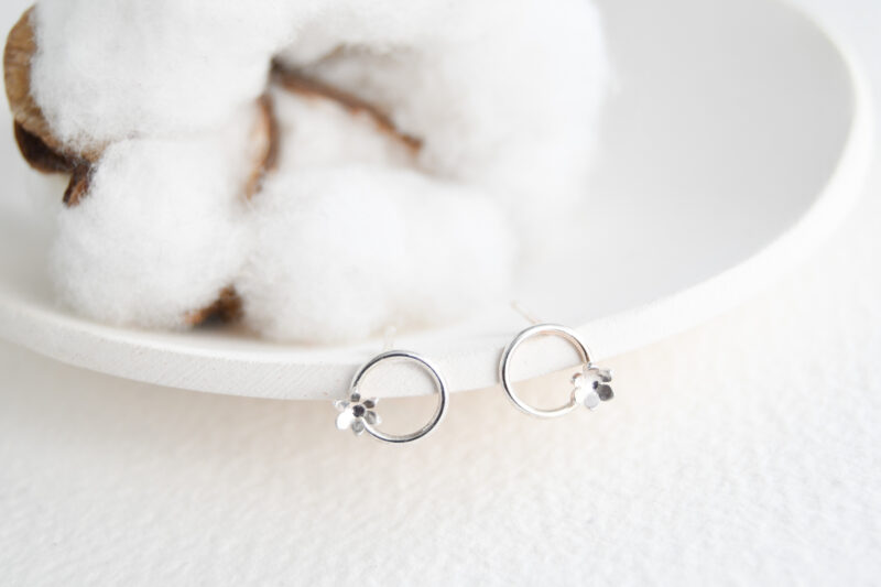White background with a white dish. Cotton flower in the background. Sterling silver studs sit on the edge of the dish. Sterling silver circle earrings with little flowers on the circle on the bottom just off centre.