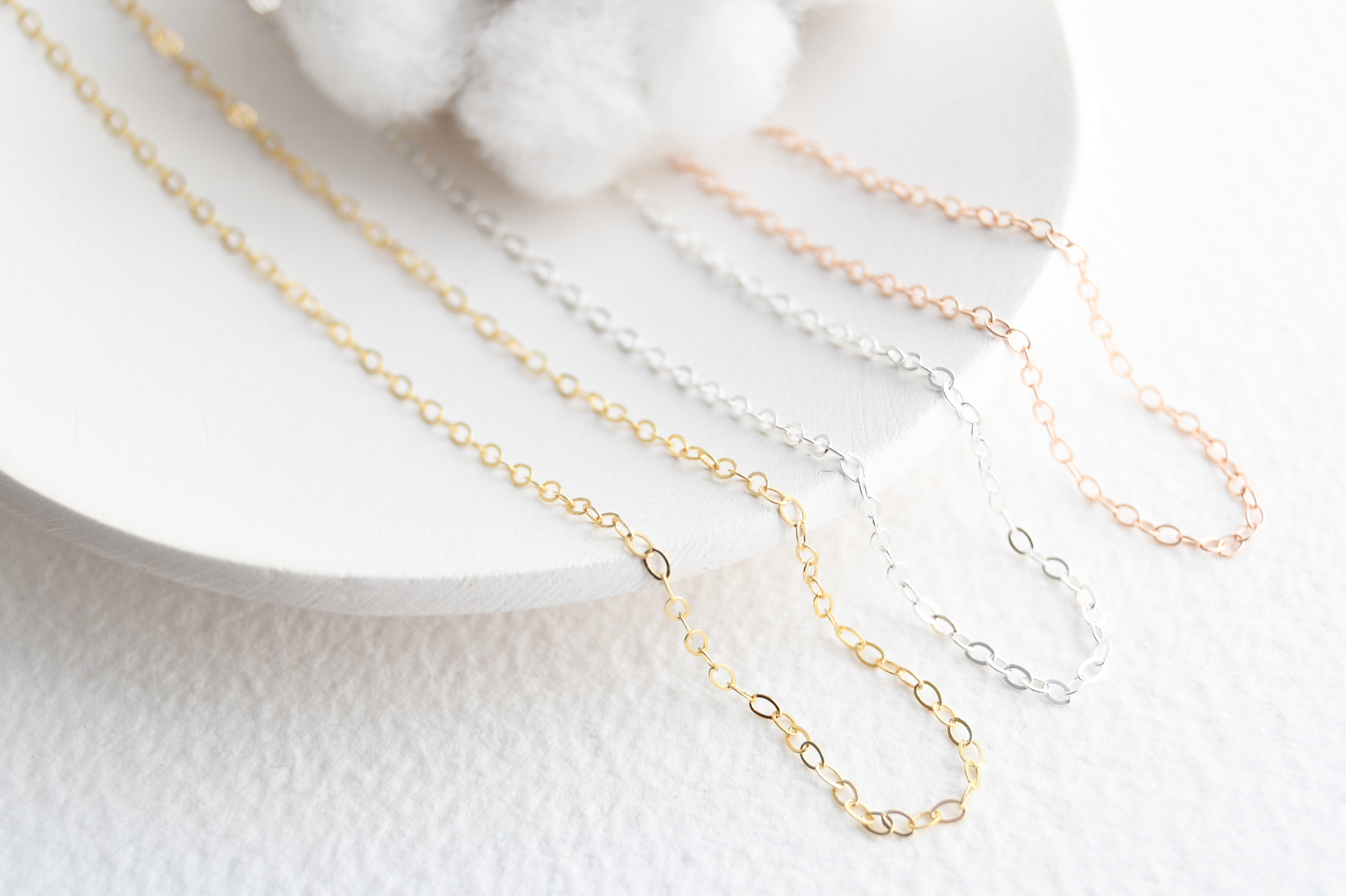 Three cable chain necklaces laying on a white dish. Necklace colours gold filled, sterling silver, and rose gold filled. A cotton flower in the background.
