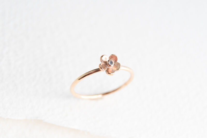 White background with rough edge paper. Gold filled ring in the middle of the picture. The ring has a 5 petal daisy flower on it with a zirconia stone, bezel set (metal covering end edge of the stone) in the middle of the flower.
