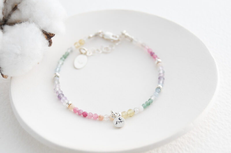 White background with a white dish in the middle of the photo. Part of a cotton flower is in the left hand corner. A multi coloured round beaded gemstone bracelet is on the white dish. 5 silver beads are in the bracelet. A small round charm with a mountain on it, is in the middle of the bracelet.