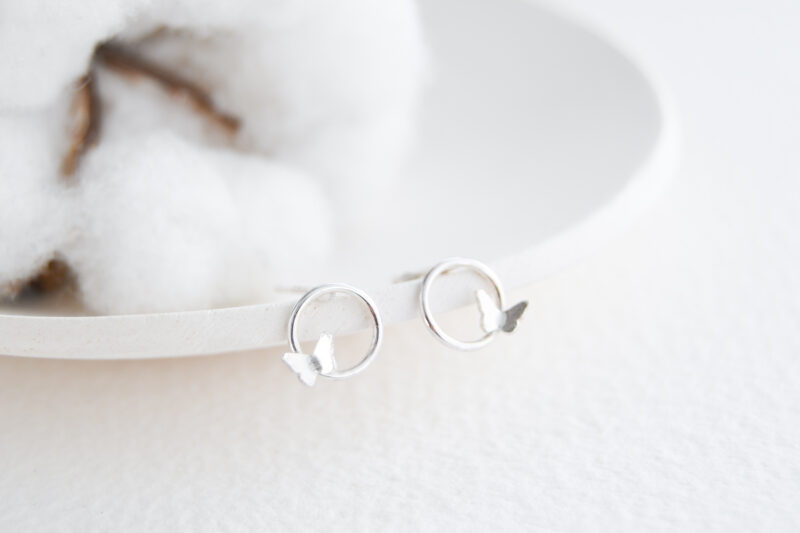 White background with a cotton flower and white ring dish. Sterling silver circle stud earrings with little butterflies sit on the edge of the ring dish.