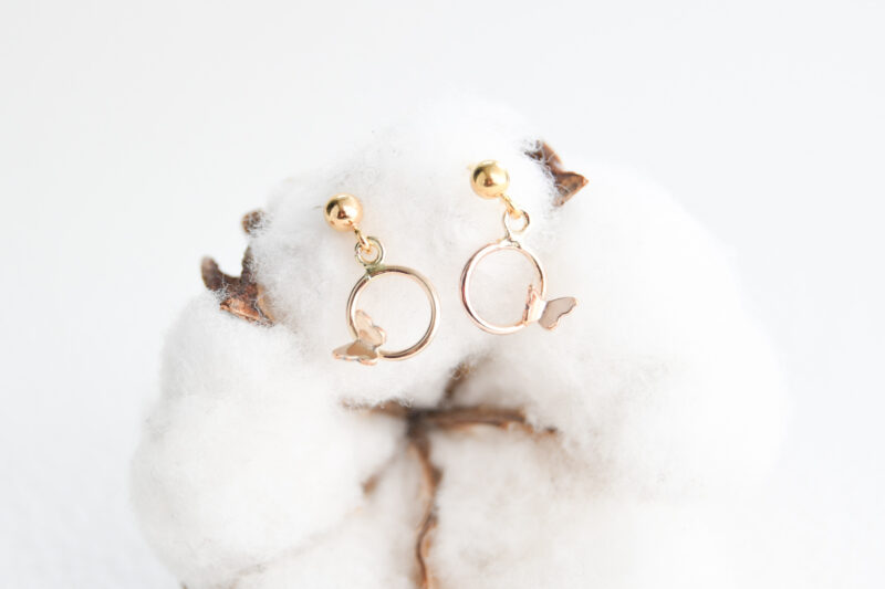 enlarged cotton flower in the background. Gold filled earrings on the cotton flower. The gold filled earrings have a ball stud at the top. Gold filled circles are attached to the ball studs with small butterflies, off centre, attached to the gold filled circles.